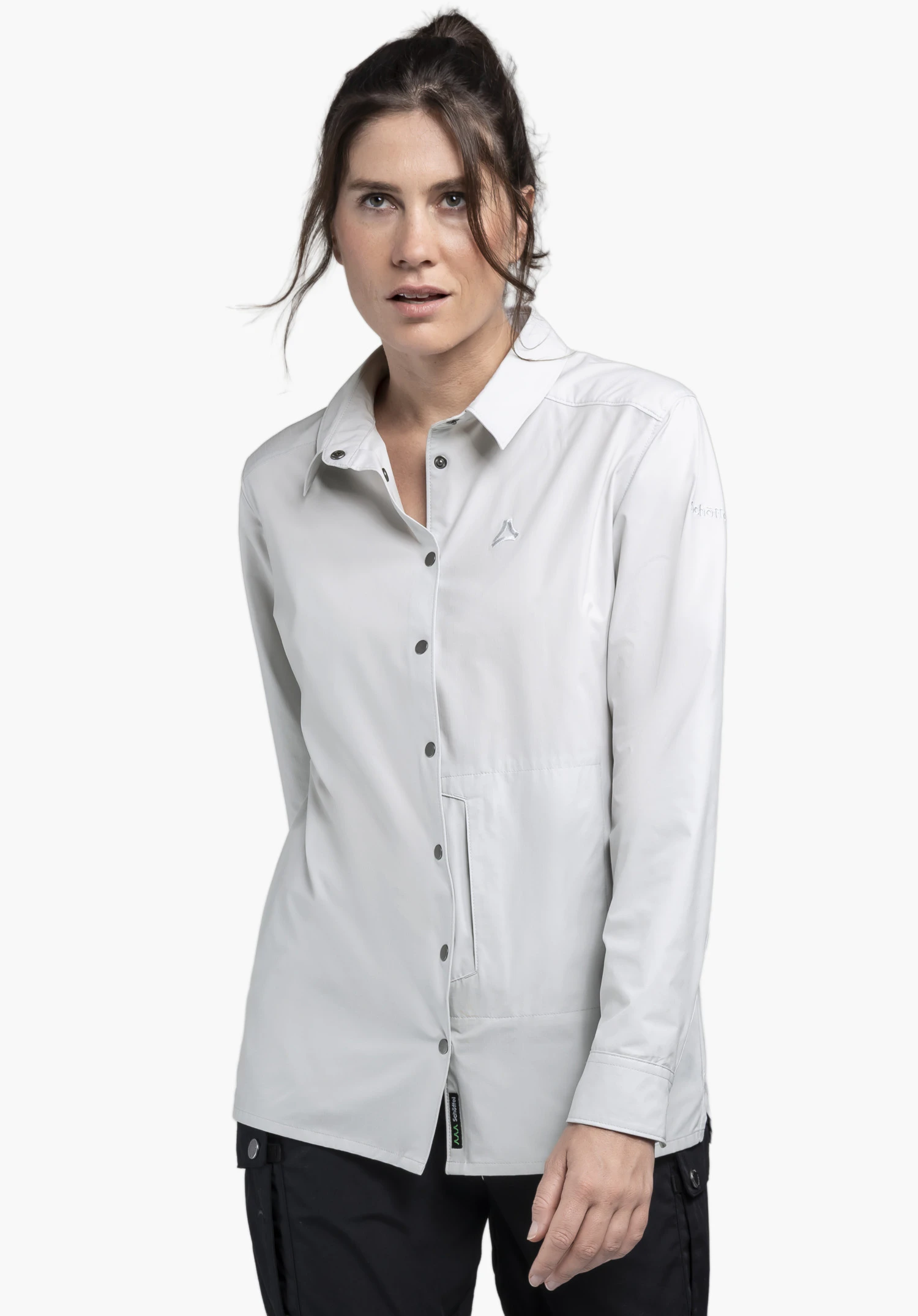 Blouse Style Maghera WMN en gris présentée sur un mannequin devant un fond neutre. Le modèle est face à la caméra et porte la blouse avec ses boutons-pression caractéristiques, tandis que le logo Schöffel discret sur la poitrine souligne le design.
