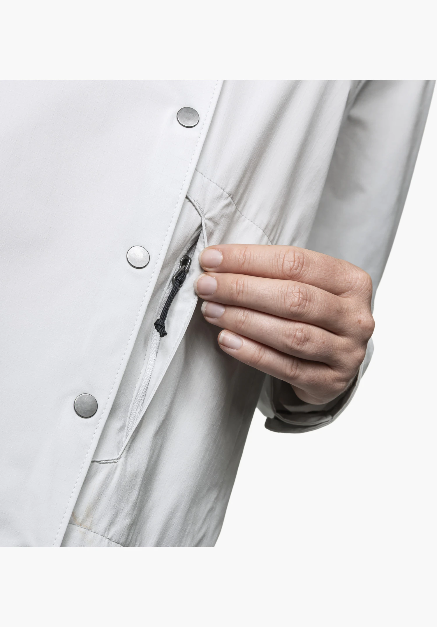Blouse Style Maghera WMN en gris, vue détaillée d'une main saisissant le petit cordon noir de la fermeture éclair de la poche latérale dissimulée, à côté d'une patte de boutonnage avec des boutons-pression argentés. Gros plan sur fond blanc/neutre.