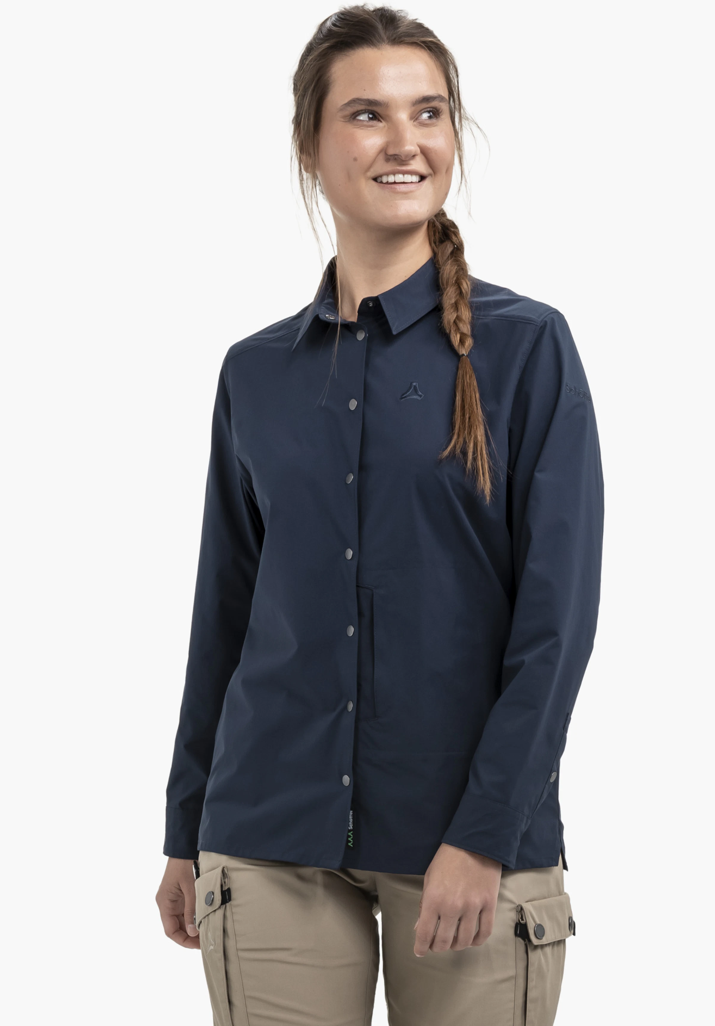 Blouse Style Maghera WMN in Blau: Eine lächelnde Frau steht halb seitlich vor einem neutralen Hintergrund. Die Bluse präsentiert sich in elegantem Blau mit dezentem Schöffel-Logo auf der Brust und am Ärmel.