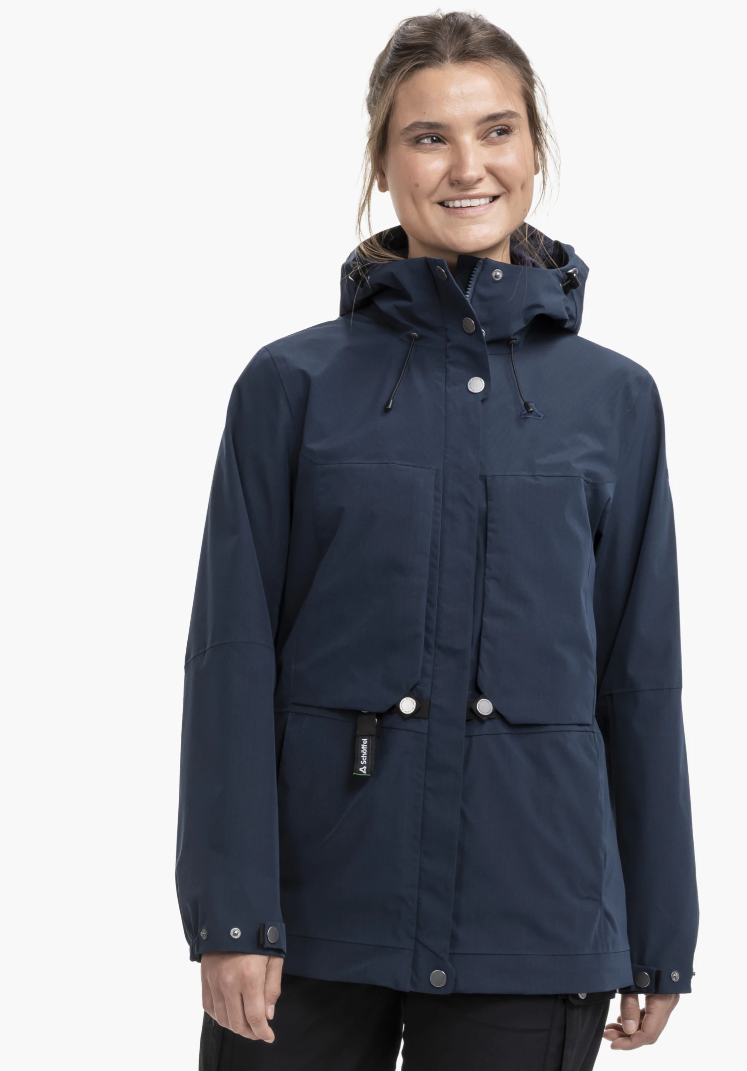 2L Jacket Style Maghera WMN : Une femme souriante porte la veste bleue sur un fond neutre. Cette vue de face met en valeur la capuche et le logo Schöffel discrètement placé sur la sangle de fermeture.