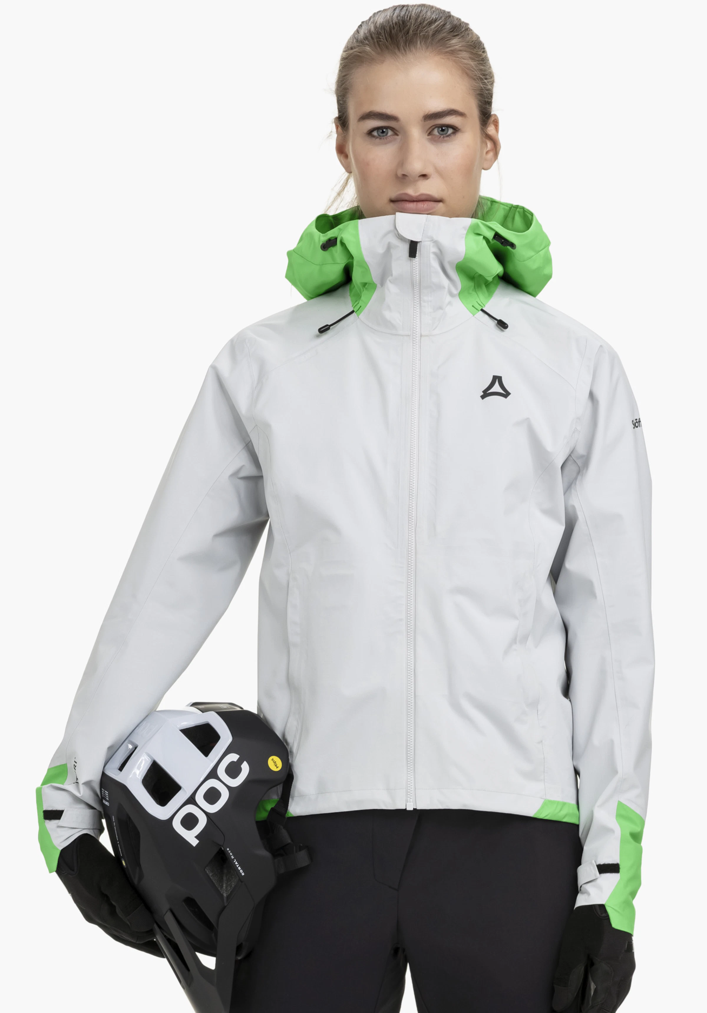 2.5L Jacket Style Tajiva WMN an einem Model, das frontal steht und einen Helm unter dem rechten Arm hält. Die Jacke in Grau zeigt ein deutlich sichtbares Schöffel-Logo auf der linken Brust und wird vor einem neutralen Hintergrund präsentiert.