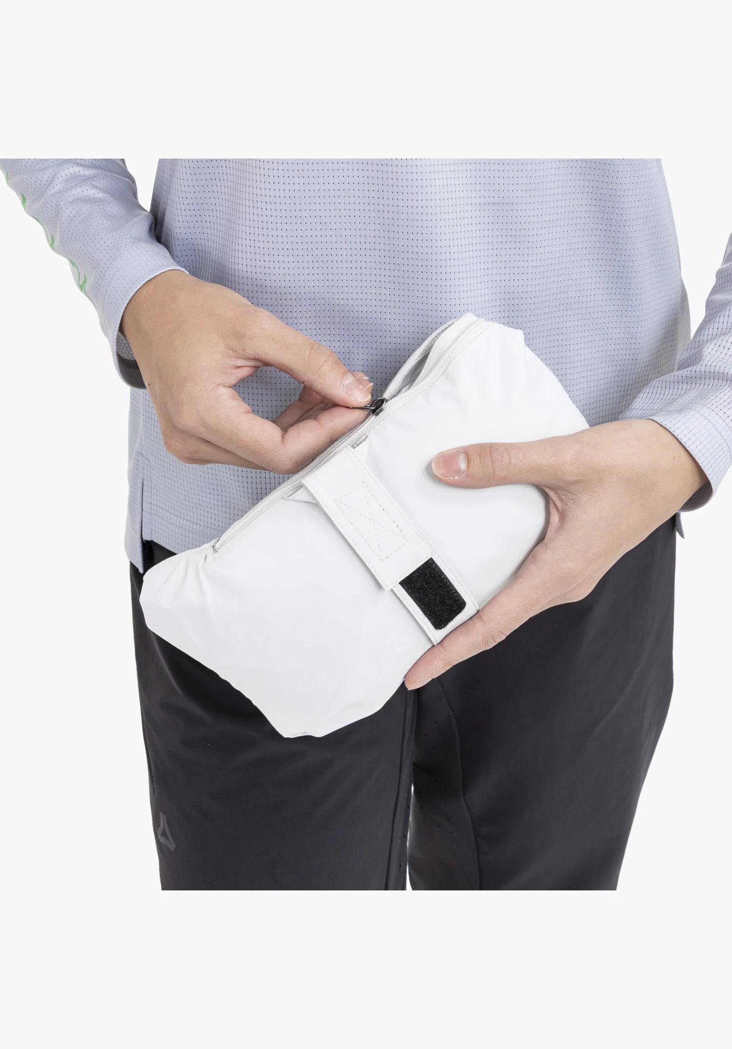 2.5L Jacket Style Tajiva WMN, détail de la veste grise rangée dans sa propre poche, montrant une fermeture zippée visible et une sangle de fixation avec bande auto-agrippante noire, tenue par deux mains. Gros plan sur fond blanc/neutre.