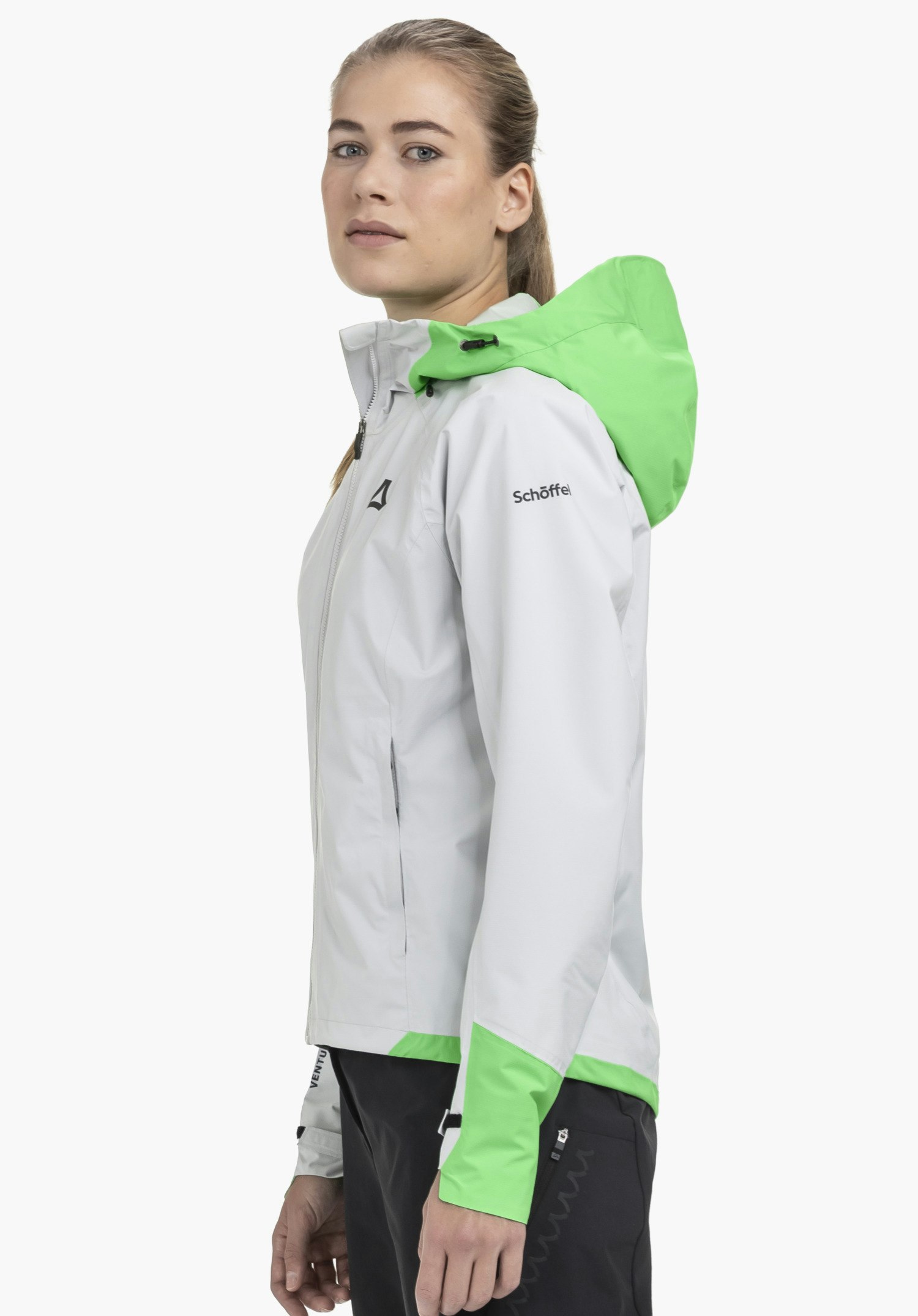 2.5L Jacket Style Tajiva WMN in grey worn by a model standing in profile against a neutral background. The jacket features contrasting green accents on the hood and cuffs, while the subtle Schöffel logo on the upper arm adds a signature brand touch.