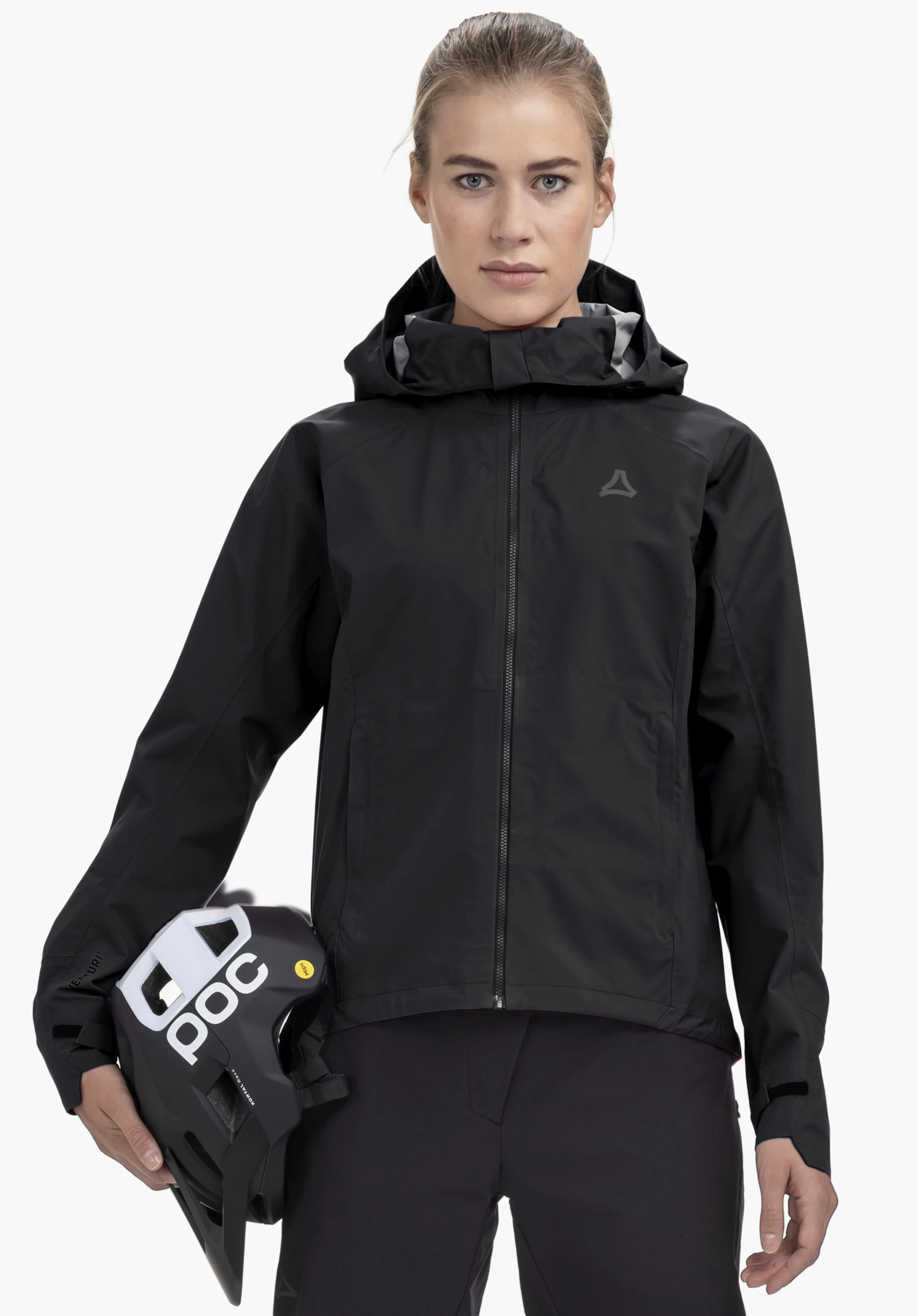 2.5L Jacket Style Tajiva WMN. Ein Model steht frontal und trägt die schwarze Jacke mit sichtbarem Schöffel-Logo auf der Brust. Sie hält einen Helm unter dem Arm, fotografiert vor einem neutralen Hintergrund.