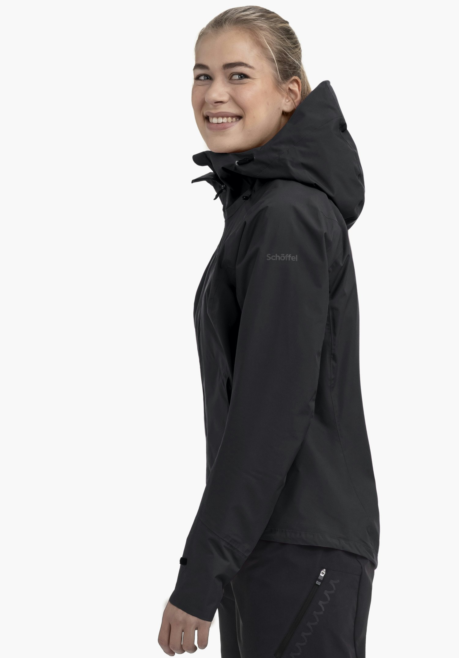 2.5L Jacket Style Tajiva WMN präsentiert in der Farbe schwarz an einem Model, das seitlich in die Kamera lächelt. Die Aufnahme vor neutralem Hintergrund hebt die Kapuze und das dezente Schöffel-Logo am Oberarm hervor.