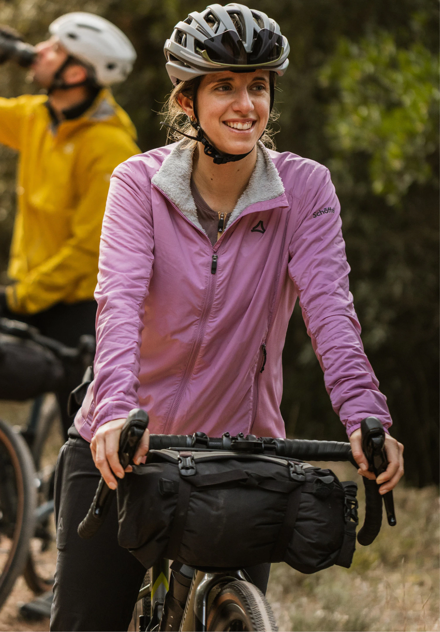 Ins Jacket Style Samaia WMN. A model stands frontally in front of a neutral background, wearing the pink jacket with a visible Schöffel-Logo on the chest whilst holding a cycling helmet in her hand.
