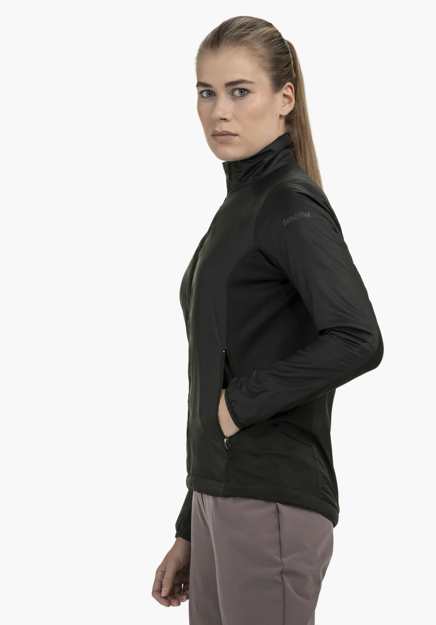 Ins Jacket Style Samaia WMN: Das Model steht seitlich vor einem neutralen Hintergrund und trägt die Jacke in der Farbe schwarz, die sich durch ihr schlichtes Design auszeichnet. Das dezente Schöffel-Logo am Oberarm unterstreicht den hochwertigen Look des Modells, während die Hand in der praktischen Tasche liegt.