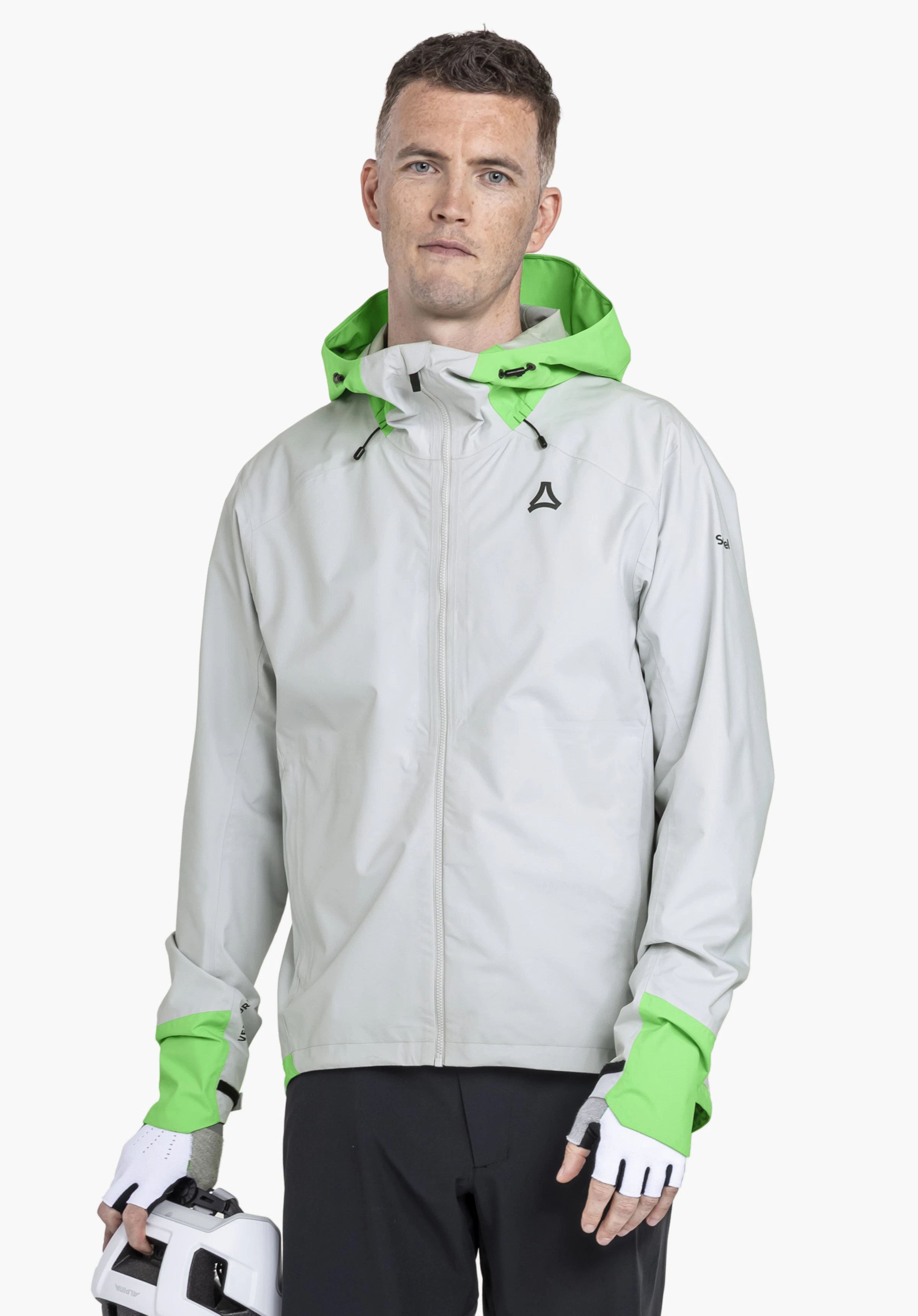 2.5L Jacket Style Tajiva MEN: Een model staat frontaal voor een neutrale achtergrond en draagt de jas in de kleur grijs. Het Schöffel-Logo is zichtbaar op de linkerborst, terwijl de man een fietshelm in zijn hand houdt.