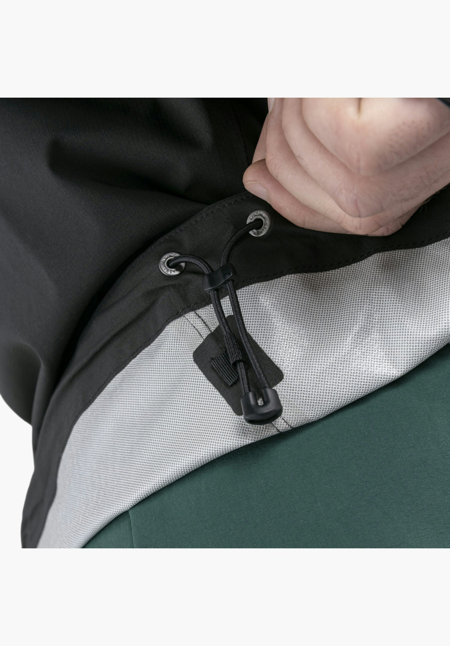 2.5L Jacket Style Tajiva MEN zeigt das Detail des verstellbaren, schwarzen Kordelzugs mit Stopper und geprägten Ösen mit Schöffel-Logo am Saum, der von einer Hand gegriffen wird und das helle Innenfutter offenbart, als Nahaufnahme vor weißem/neutralem Hintergrund.