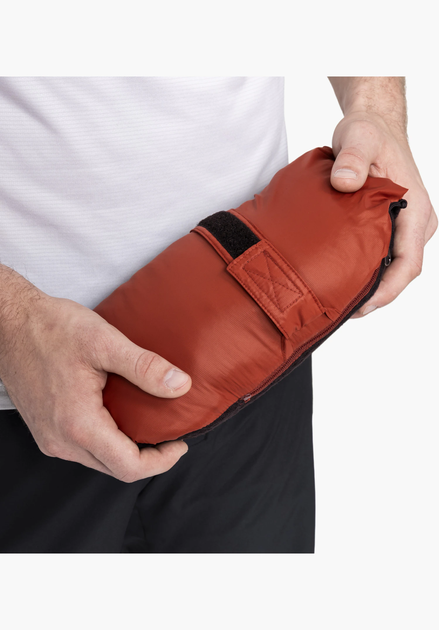 Ins Jacket Style Samaia MEN en rouge, repliée en un paquet compact tenu entre les mains, avec un accent sur le tissu lisse et la sangle de fermeture auto-agrippante fonctionnelle. Gros plan sur fond blanc/neutre.