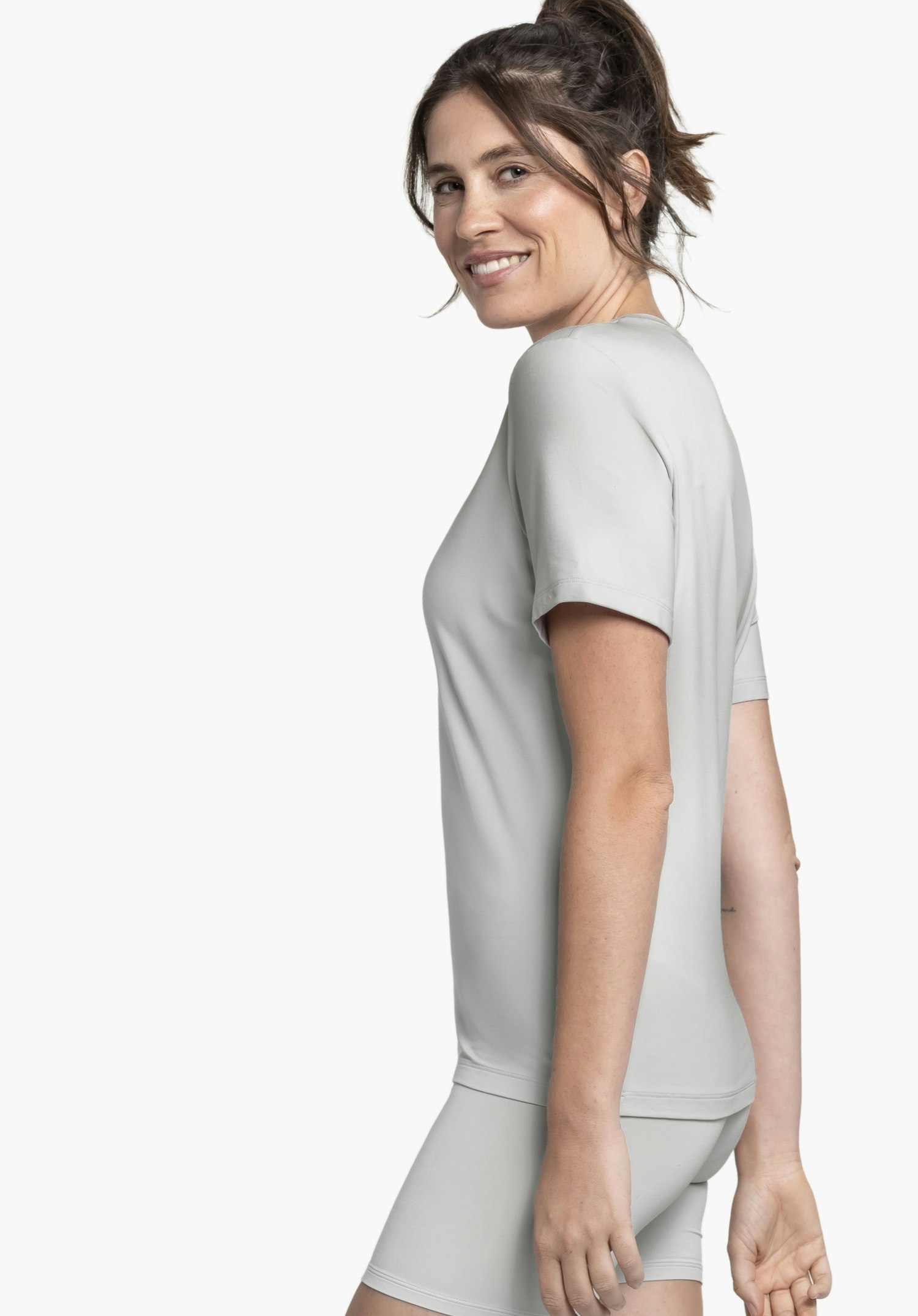 T Shirt Style Mafadi WMN en gris : Un mannequin souriant se tient de profil devant un fond neutre pour présenter ce t-shirt. La coupe épurée est mise en valeur par cette vue latérale, le design minimaliste ne présente pas de logo Schöffel visible.