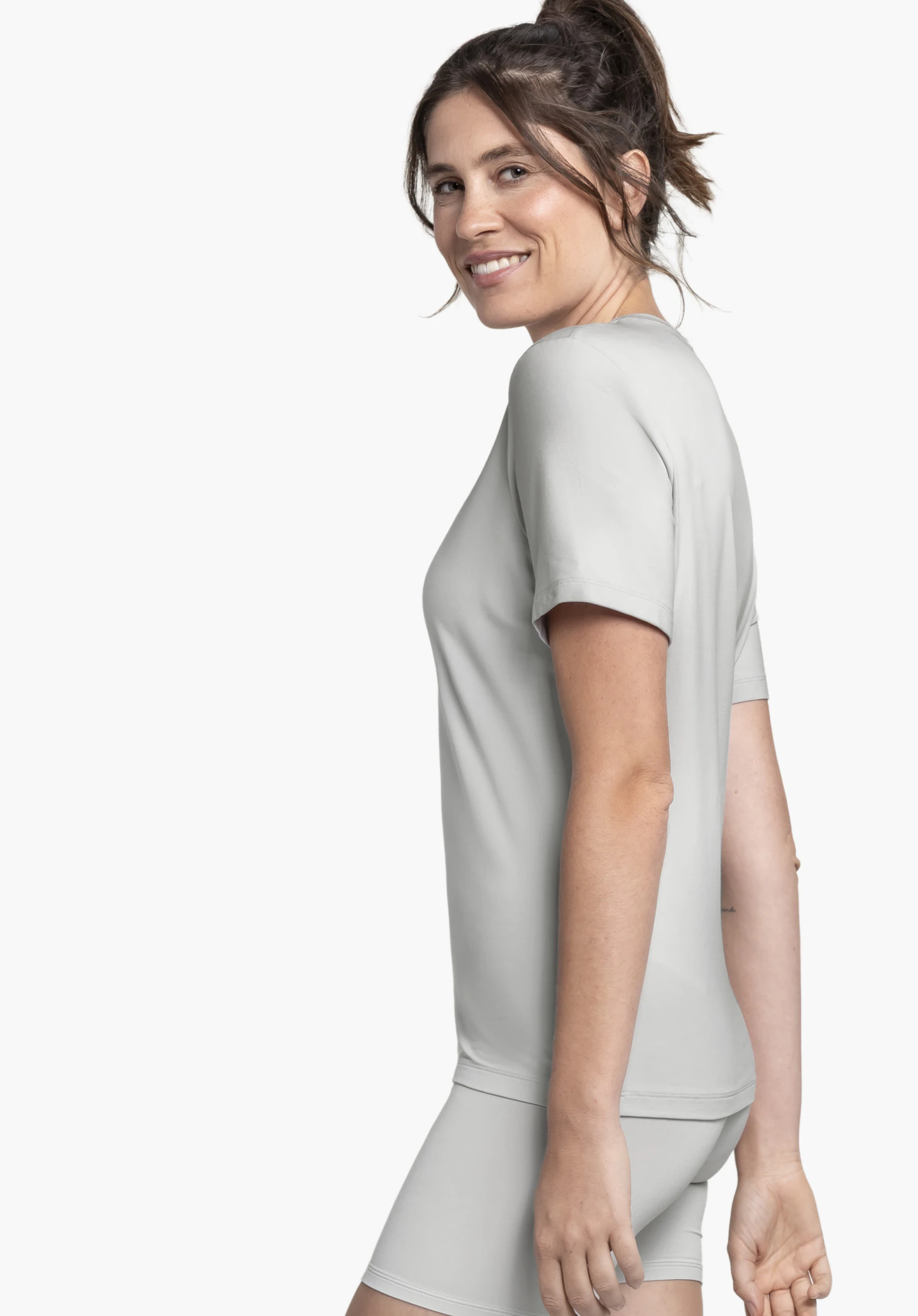 T Shirt Style Mafadi WMN en gris : Un mannequin souriant se tient de profil devant un fond neutre pour présenter ce t-shirt. La coupe épurée est mise en valeur par cette vue latérale, le design minimaliste ne présente pas de logo Schöffel visible.