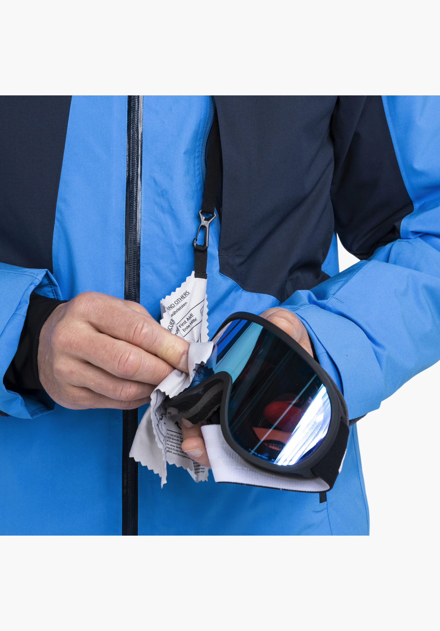 Jacket Style Trifex MEN in blue featuring an integrated goggle cleaning cloth with a carabiner and "First Aid!" print while cleaning ski goggles in detail, close-up shot against a white background.