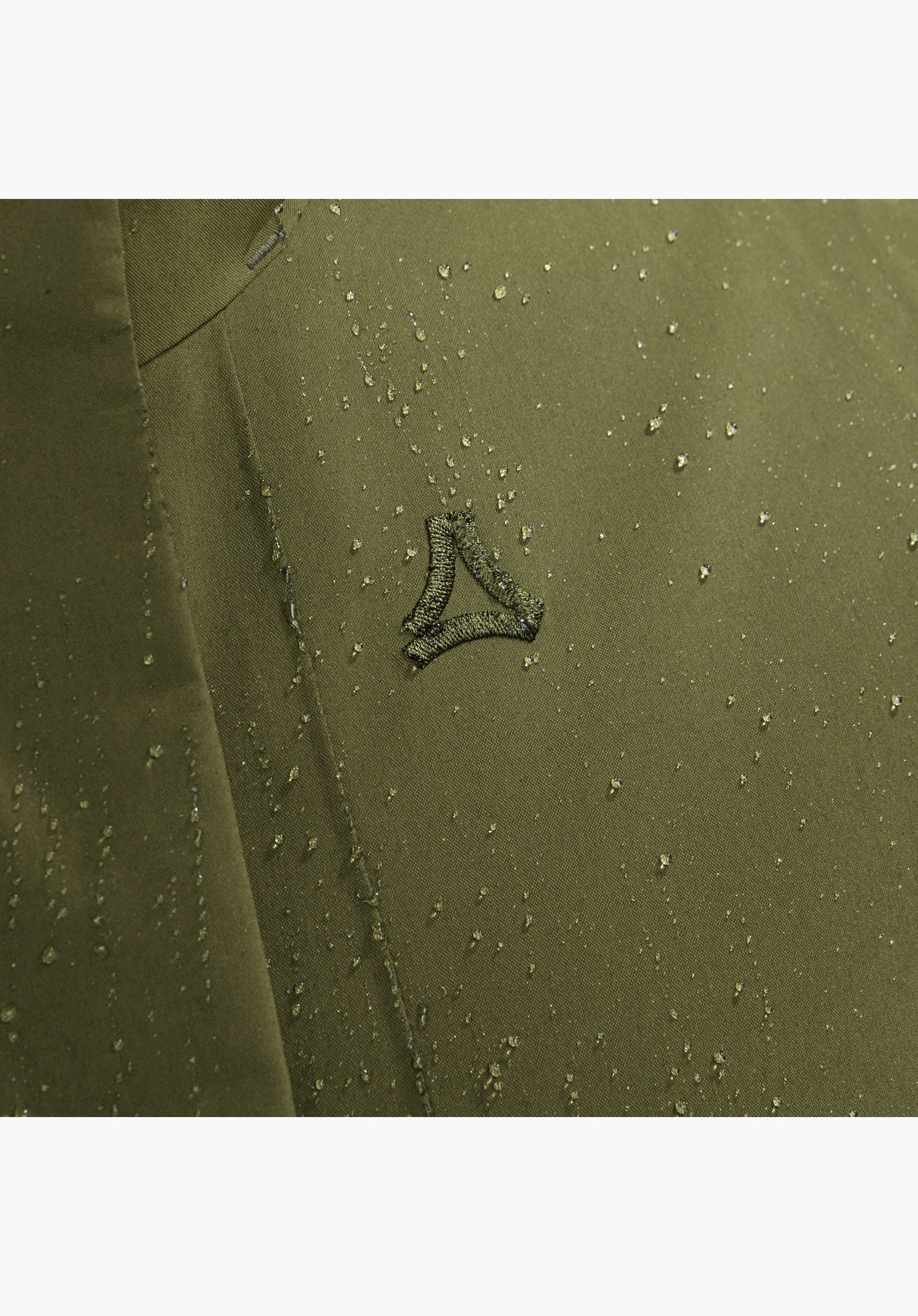 Ins Coat Style Optimea WMN gros plan sur la matière verte avec le logo Schöffel brodé et des gouttes d'eau perlantes en gros plan sur fond neutre.