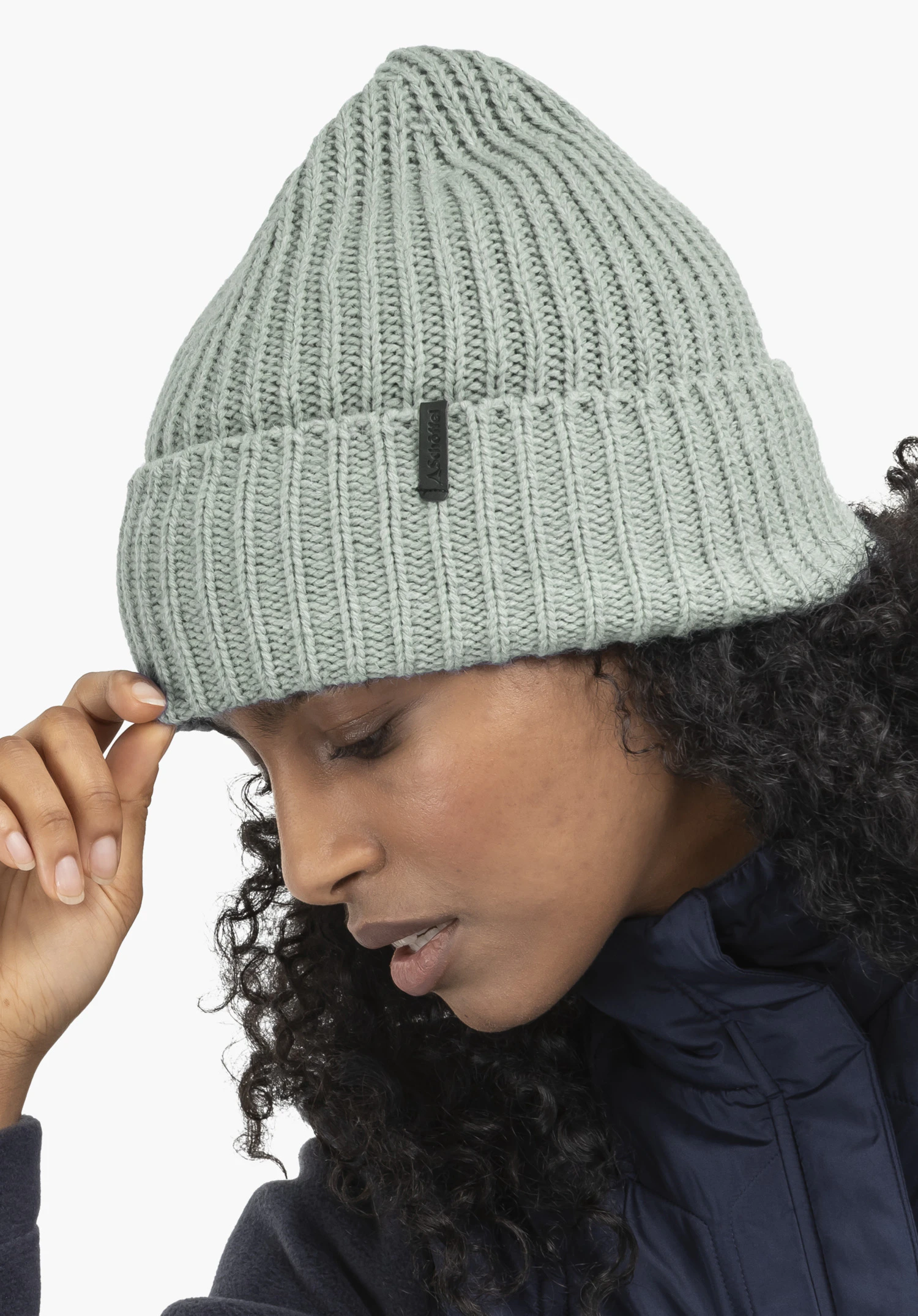 Knitted Hat Medford in grey with Schöffel lettering, worn by a female model in a side view gently touching the beanie, against a white background. (AI-generated)